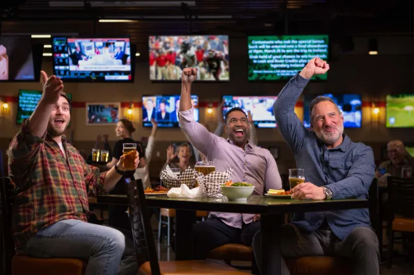 3 men cheering at a table with food and beer with TVs playing in the background