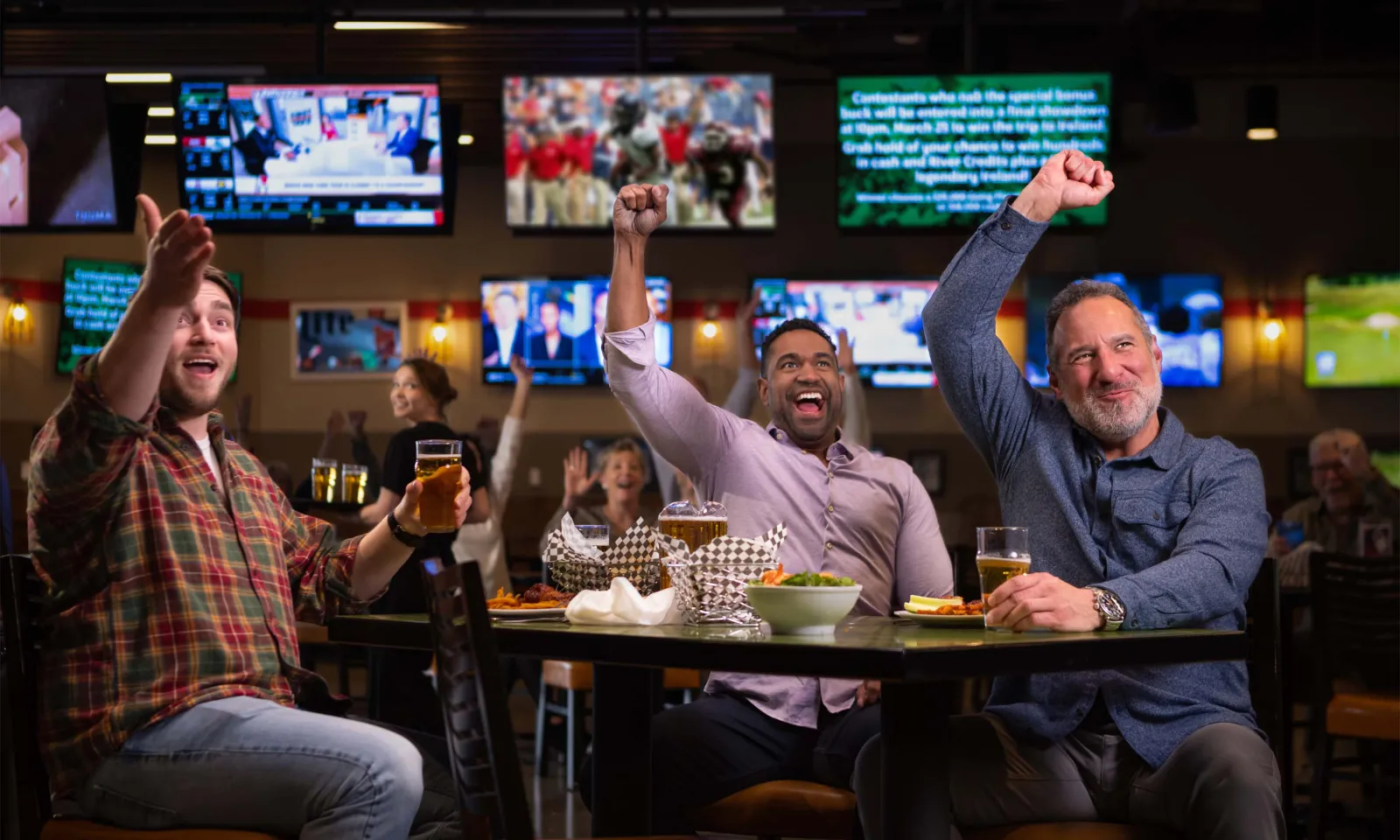 3 men cheering at a table with food and beer with TVs playing in the background